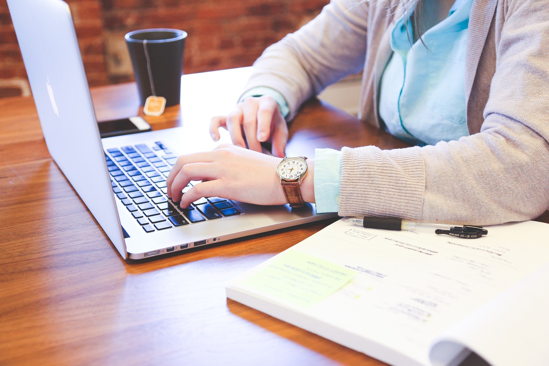 Worker sitting at a desk with laptop, drinking a mug of tea and writing in a book
